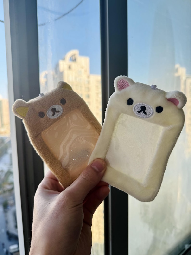 May include: Two plush bear-shaped ID badge holders. One is tan, the other is off-white. Both have a clear plastic window for an ID card, embroidered black eyes, and a small black nose. The ears have pink accents.
