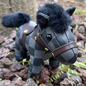 May include: A stuffed horse figurine with a black and grey chequered pattern. The horse has a black fur mane and tail, a brown saddle and bridle, and gold-coloured accents. The toy is set on a bed of small rocks and leaves.