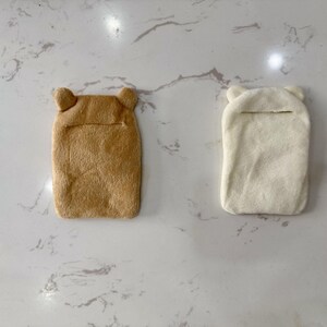 May include: Two plush, rectangular hot water bottle covers. One is tan, the other is off-white. Both have bear-shaped ears at the top and a slit opening. The covers are designed to hold a hot water bottle.