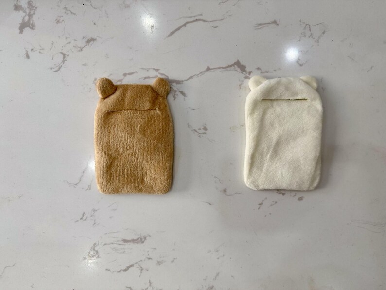 May include: Two plush, rectangular hot water bottle covers. One is tan, the other is off-white. Both have bear-shaped ears at the top and a slit opening. The covers are designed to hold a hot water bottle.