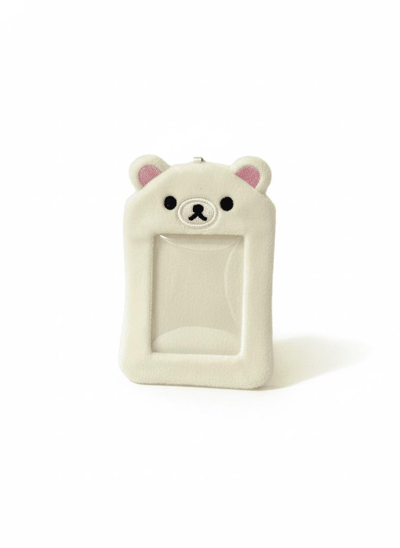 May include: A white, plush bear-shaped ID holder with a clear window. The bear has pink ears, black eyes, and a stitched black nose and mouth. The holder is rectangular with rounded edges and a small loop at the top.