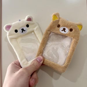 May include: Two plush bear-shaped ID badge holders. One is cream-colored with pink ears, the other is tan with yellow ears. Both have clear plastic windows for displaying an ID. The holders are being held by a hand.