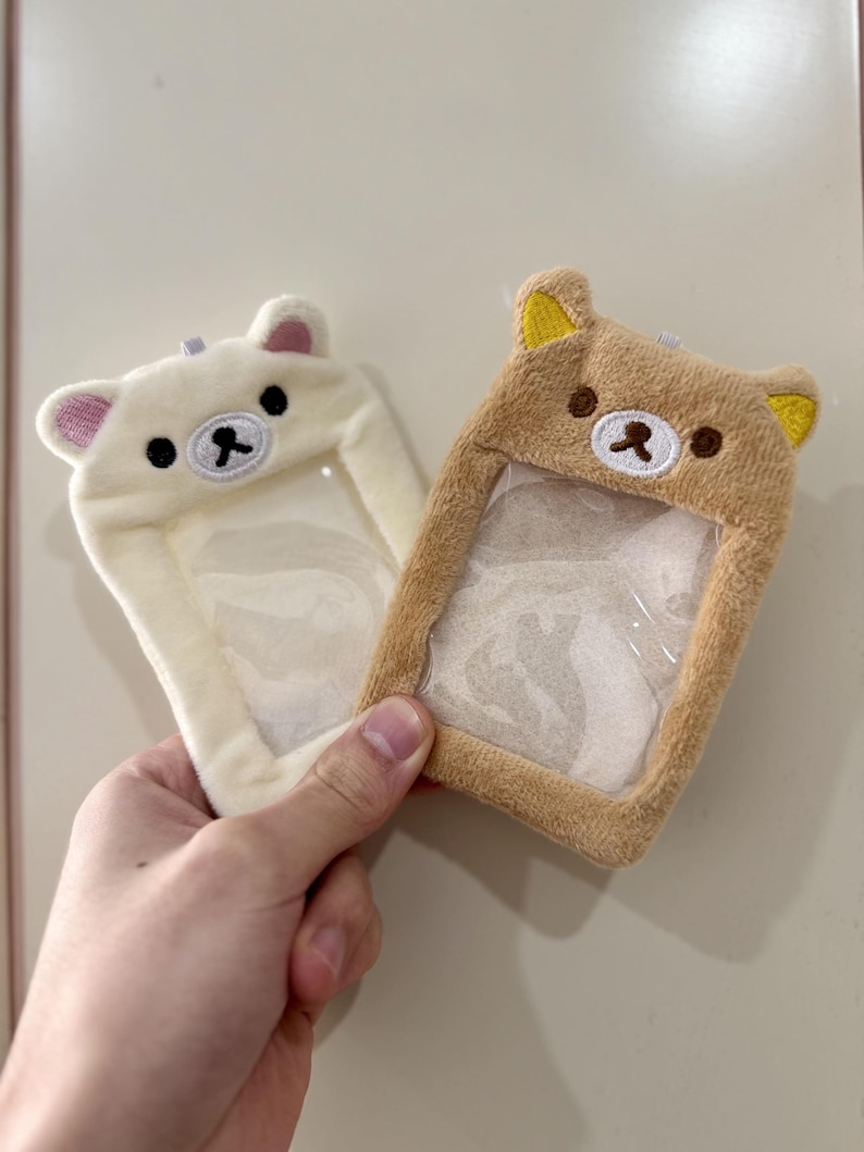 May include: Two plush bear-shaped ID badge holders. One is cream-colored with pink ears, the other is tan with yellow ears. Both have clear plastic windows for displaying an ID. The holders are being held by a hand.