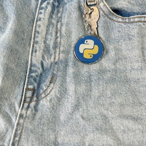 Puede incluir: Un llavero con el logotipo de Python en azul y amarillo, sujeto a unos vaqueros de mezclilla de lavado claro. El llavero es redondo con un borde plateado, y tambi&eacute;n hay una llave adjunta. Los vaqueros tienen un dise&ntilde;o cl&aacute;sico de cinco bolsillos.
