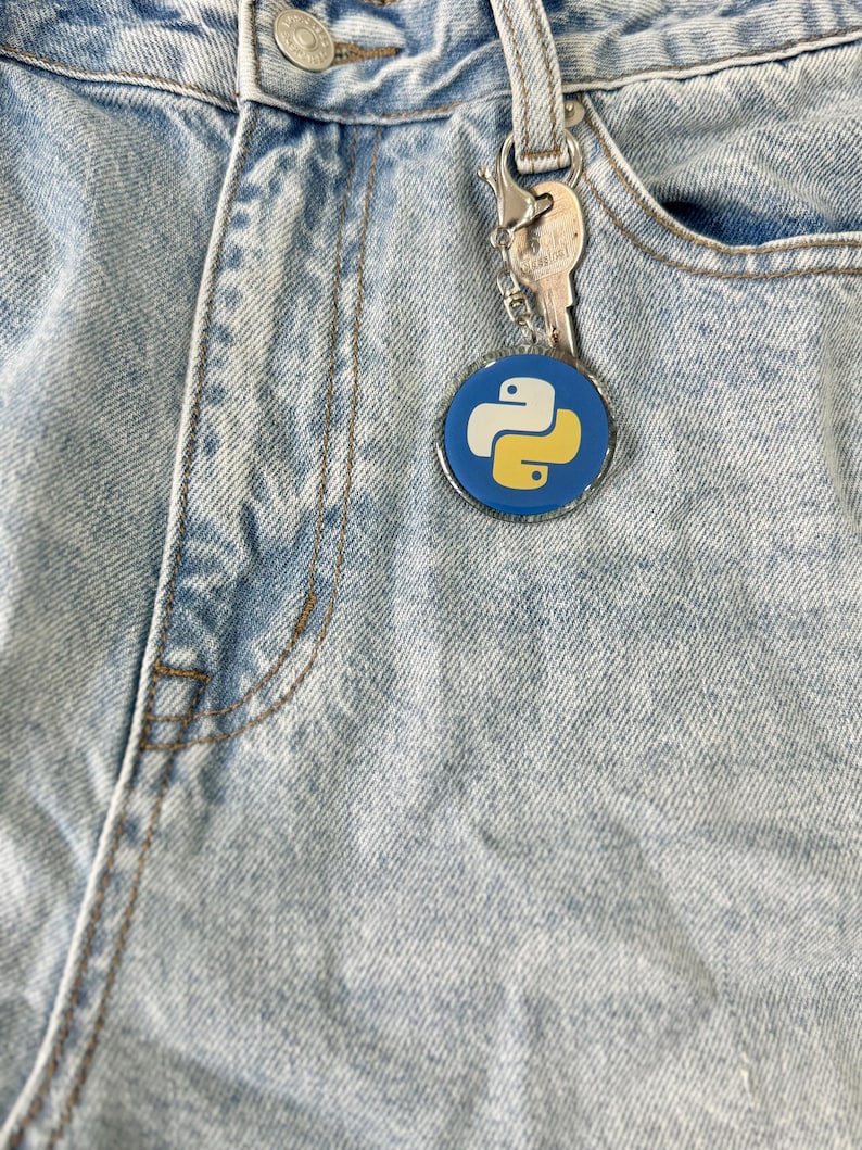 Puede incluir: Un llavero con el logotipo de Python en azul y amarillo, sujeto a unos vaqueros de mezclilla de lavado claro. El llavero es redondo con un borde plateado, y tambi&eacute;n hay una llave adjunta. Los vaqueros tienen un dise&ntilde;o cl&aacute;sico de cinco bolsillos.