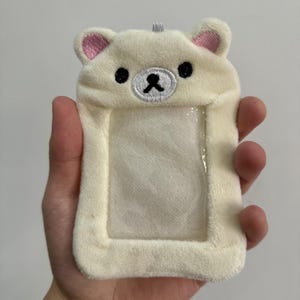 May include: A plush, off-white ID holder shaped like a bear's head, featuring pink inner ears, black eyes, and a black nose. The holder has a clear plastic window for displaying an ID or card. A small loop is at the top for attaching a lanyard.