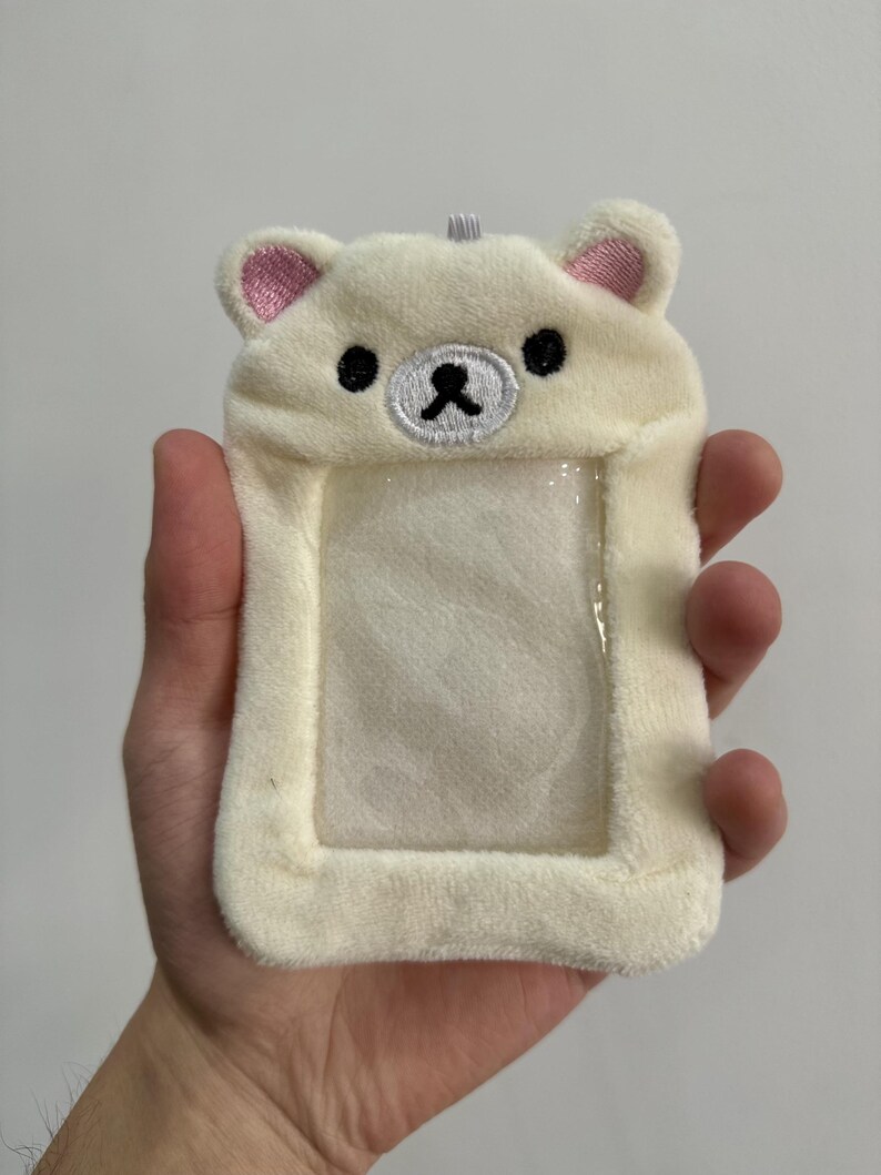 May include: A plush, off-white ID holder shaped like a bear's head, featuring pink inner ears, black eyes, and a black nose. The holder has a clear plastic window for displaying an ID or card. A small loop is at the top for attaching a lanyard.