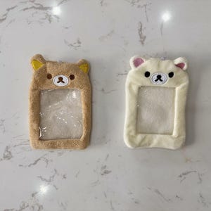 May include: Two plush bear-shaped ID badge holders. One is tan with yellow ears, the other is white with pink ears. Both have a clear plastic window for displaying an ID card. The bears have embroidered facial features.