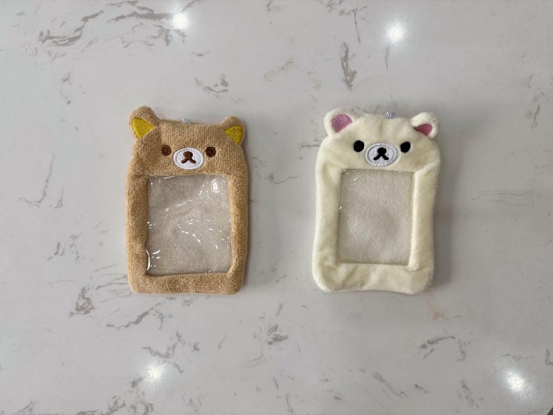 May include: Two plush bear-shaped ID badge holders. One is tan with yellow ears, the other is white with pink ears. Both have a clear plastic window for displaying an ID card. The bears have embroidered facial features.