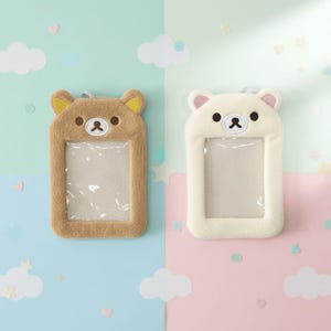 May include: Two plush bear-shaped ID badge holders. One is tan with yellow ears, the other is white with pink ears. Both have a clear plastic window for displaying an ID. The background is split between light blue and pink.