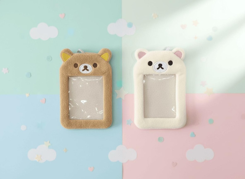 May include: Two plush bear-shaped ID badge holders. One is tan with yellow ears, the other is white with pink ears. Both have a clear plastic window for displaying an ID. The background is split between light blue and pink.