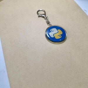 Puede incluir: Un llavero redondo con fondo azul y el logotipo de Python en blanco y amarillo. El llavero tiene un borde met&aacute;lico plateado y un cierre para sujetarlo a llaves o bolsos.