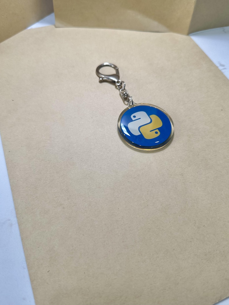 Puede incluir: Un llavero redondo con fondo azul y el logotipo de Python en blanco y amarillo. El llavero tiene un borde met&aacute;lico plateado y un cierre para sujetarlo a llaves o bolsos.