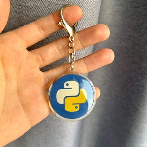 Puede incluir: Un llavero redondo con fondo azul y el logotipo de Python en blanco y amarillo. El llavero tiene un cierre y una cadena plateados. El dise&ntilde;o es un s&iacute;mbolo popular de la programaci&oacute;n inform&aacute;tica.