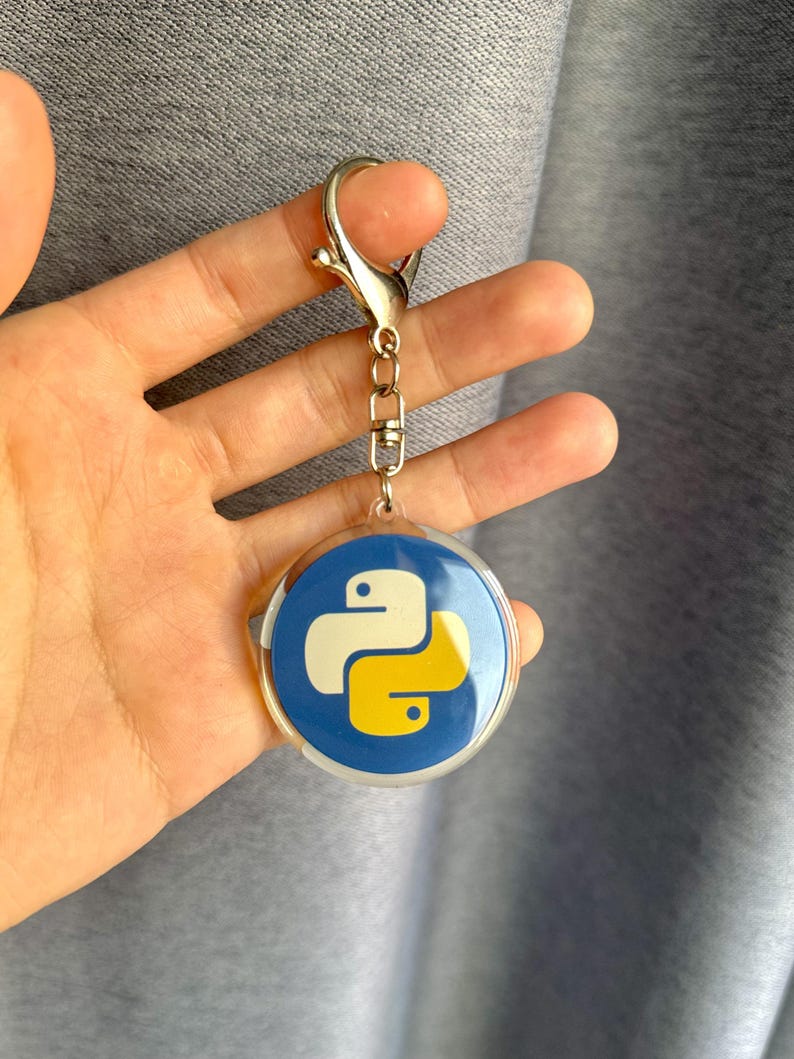 Puede incluir: Un llavero redondo con fondo azul y el logotipo de Python en blanco y amarillo. El llavero tiene un cierre y una cadena plateados. El dise&ntilde;o es un s&iacute;mbolo popular de la programaci&oacute;n inform&aacute;tica.