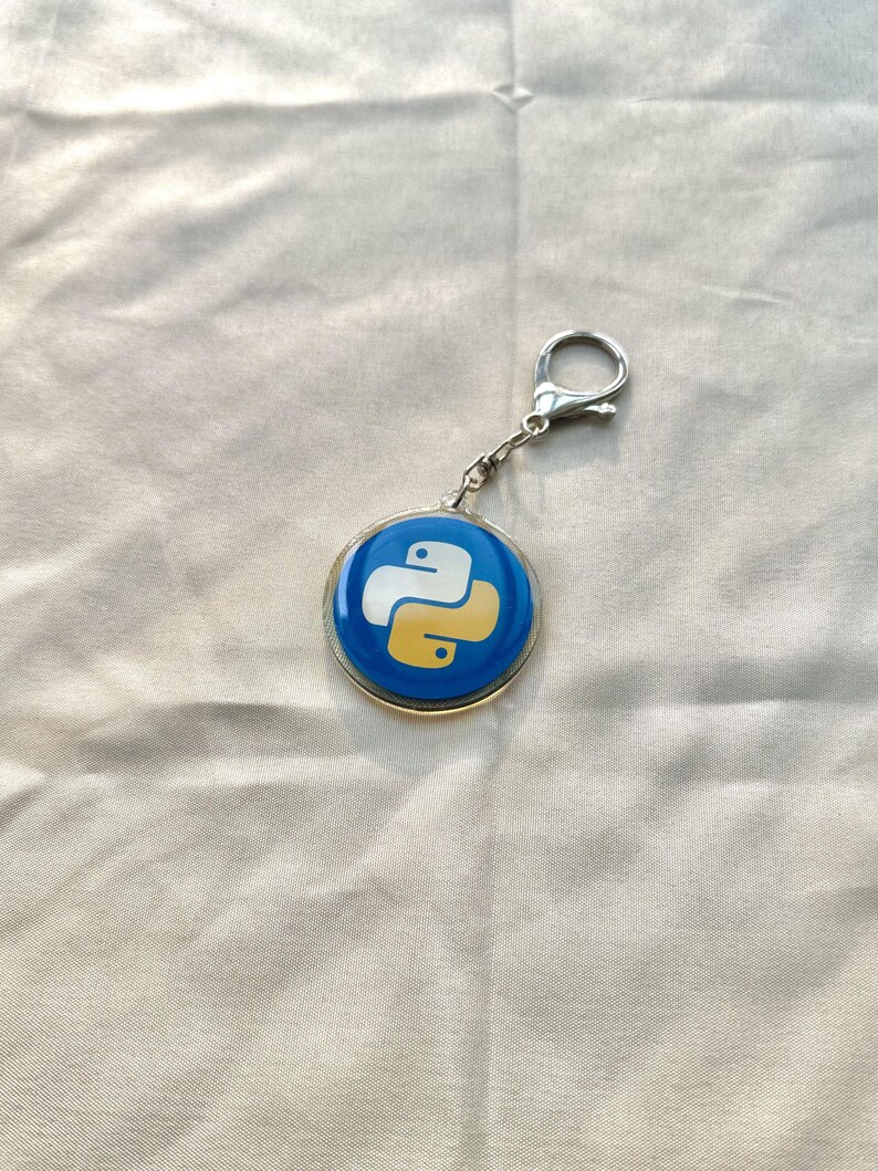 Puede incluir: Un llavero redondo con fondo azul y el logotipo de Python en blanco y amarillo. El llavero tiene un borde de metal plateado y un cierre para sujetarlo a llaves o bolsos.