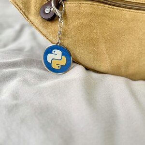 Puede incluir: Un llavero redondo con fondo azul y logotipo blanco y amarillo. El llavero est&aacute; sujeto a un cierre plateado y a un bot&oacute;n de cuero marr&oacute;n. Una ri&ntilde;onera beige est&aacute; en el fondo.