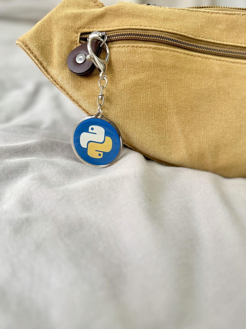 Puede incluir: Un llavero redondo con fondo azul y logotipo blanco y amarillo. El llavero est&aacute; sujeto a un cierre plateado y a un bot&oacute;n de cuero marr&oacute;n. Una ri&ntilde;onera beige est&aacute; en el fondo.