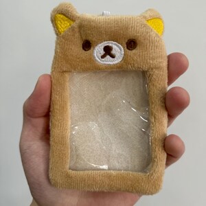 May include: A plush, light brown ID holder shaped like a bear's face. The bear has yellow ears, brown eyes, and a white nose. The holder has a clear plastic window for displaying an ID card. The item is held in a hand.