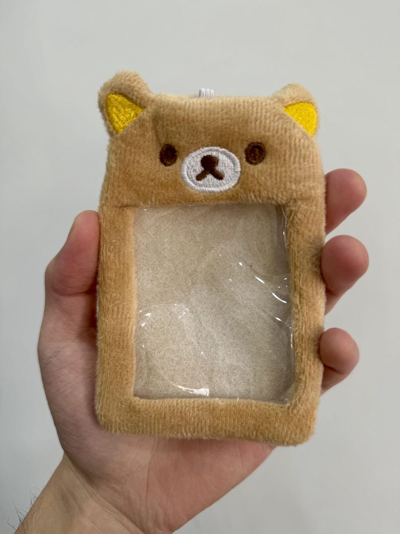 May include: A plush, light brown ID holder shaped like a bear's face. The bear has yellow ears, brown eyes, and a white nose. The holder has a clear plastic window for displaying an ID card. The item is held in a hand.