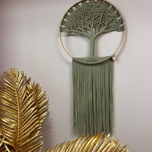 Op de afbeelding: Een saliegroene macramé wandhanger in de vorm van een boom, met een cirkelvormige gouden metalen lijst. Het boomontwerp heeft ingewikkelde knopen en de onderkant heeft lange franjes. Goudkleurige palmbladeren staan op de voorgrond.