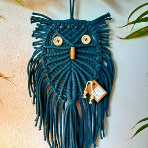 Op de afbeelding: Een teal macramé uil wandhanger. De uil heeft knoopogen en een houten snavel. Lange franjes hangen van de onderkant. Een label met de tekst "BY ME!" is bevestigd.