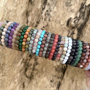 May include: A collection of beaded bracelets in various colors, including green, blue, red, and white. The bracelets are made of different types of stones and are displayed on a pink cylinder with a gold band.