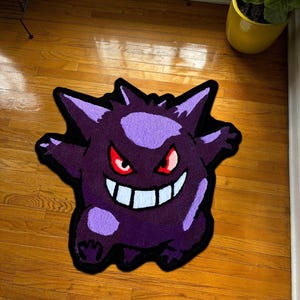Könnte beinhalten: Ein getufteter Teppich in Form der Figur Gengar, mit einem tiefvioletten Körper und hellvioletten Akzenten an Stirn und Gliedmaßen. Das Design umfasst spitze Ohren, ein breites weißes Grinsen und stechende rote Augen mit schwarzer Umrandung.