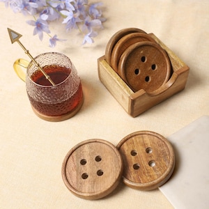 Può includere: Sottobicchieri in legno a forma di bottone con quattro fori, esposti con una tazza di tè in vetro e un cucchiaio decorativo. I sottobicchieri sono in un supporto di legno. Il set include un cucchiaio decorativo.