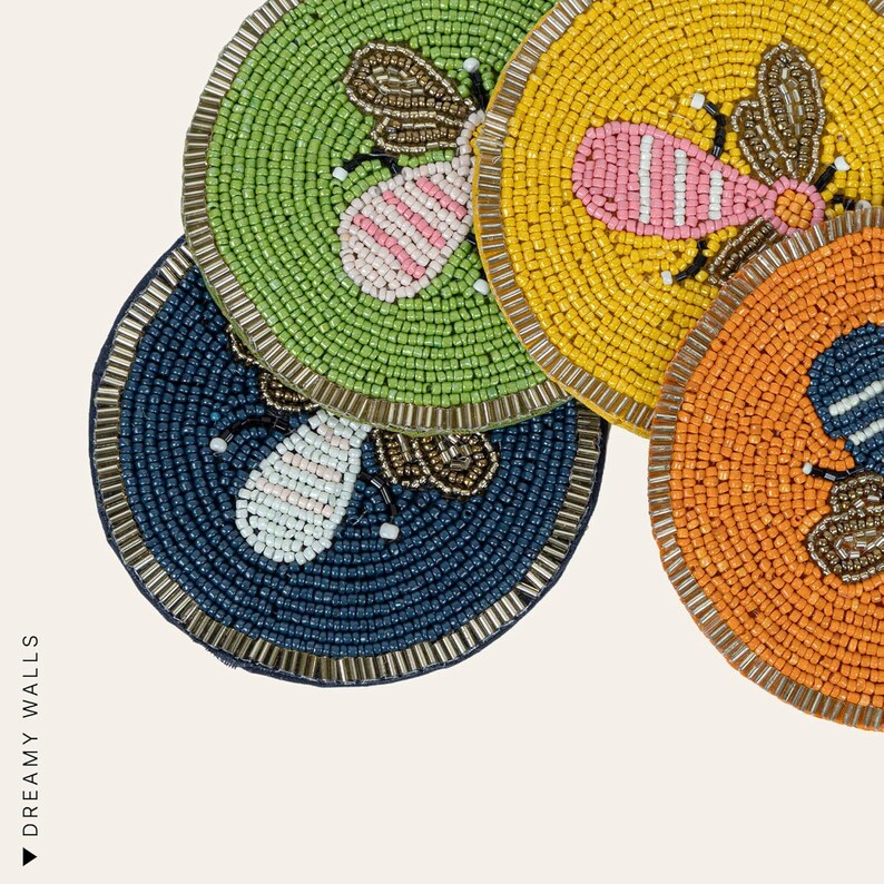 May include: Four beaded coasters in shades of green, yellow, orange, and navy blue. Each coaster features a beaded bee design with pink and white accents. The coasters have a metallic gold border and are arranged on a white background.