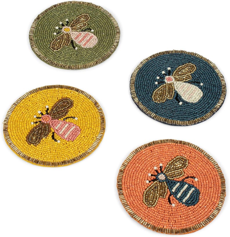 May include: Four beaded coasters with bee designs. The coasters are round and feature bees in shades of brown, pink, and white. The backgrounds are yellow, green, blue, and orange, with a gold border.