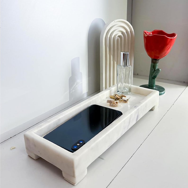 May include: A white marble tray with a rectangular shape and small feet, holding a black smartphone. Gold jewellery and a clear perfume bottle sit on the tray. A red tulip-shaped vase and a white arched decor piece are in the background.