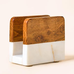 May include: A rectangular napkin holder featuring a two-tone design. The upper section is constructed from brown wood with a visible natural grain, while the lower section is made of smooth white marble with subtle grey veining throughout the base.