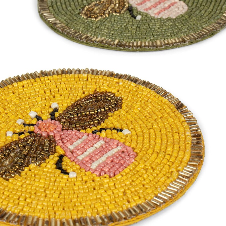 May include: Two beaded coasters with bee designs. One coaster is yellow with a brown, pink, and white bee. The other is green with a gold and pink bee. Both coasters have a gold border.