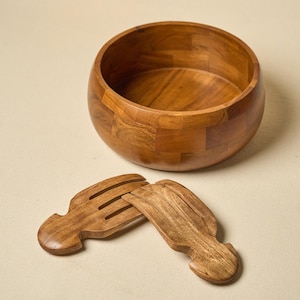 May include: A round, medium-brown wooden bowl with a visible block-patterned grain construction, accompanied by two matching wooden salad serving utensils featuring wide, slotted heads and curved handles, all displaying a smooth, polished finish.