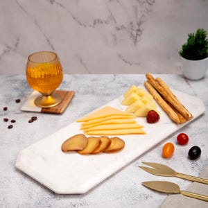 May include: A white marble serving board with various cheeses, breadsticks, and a strawberry. A glass of amber liquid sits on a wooden coaster. Gold cheese knives and a small potted plant are also present.
