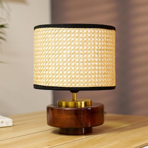 May include: A small, round table lamp with a woven, light-yellow lampshade. The lampshade is trimmed with black and sits atop a dark brown, wooden base with a brass-colored stem. The lamp is on a wooden surface.