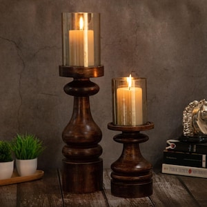 Puede incluir: Dos candelabros de madera marrón oscuro con velas encendidas dentro de cilindros de vidrio. Los candelabros tienen un diseño torneado. Las velas de color marfil están encendidas con llamas brillantes.