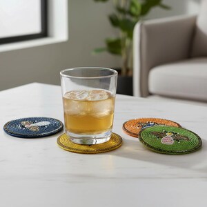 May include: A glass of amber liquid with ice cubes sits on a yellow beaded coaster. Three additional beaded coasters in blue, orange, and green are nearby. Each coaster features a beaded insect design.