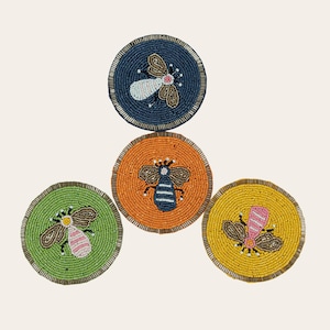 May include: A set of beaded bee coasters. The coasters are circular with a beaded edge. Each coaster has a different background colour: navy, orange, green, and yellow. The bees are also different colours, including white, brown, and pink.