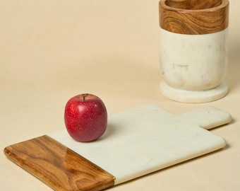May include: A rectangular white marble serving board with a polished wooden handle and base edge, paired with a matching cylindrical marble and wood utensil holder. A single bright red apple rests on the surface of the serving board.