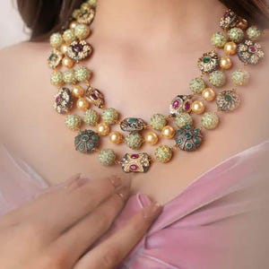 May include: A multi-strand necklace with gold-coloured beads and ornate, jewel-encrusted pendants. The necklace features green, red, and gold gemstones. The jewellery is displayed on a person wearing a pink garment.