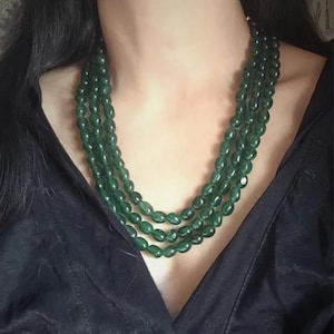 May include: A multi-strand necklace featuring dark green oval beads. The necklace is draped around the neck, showcasing the rich color and texture of the beads. The background is a dark, solid color, which highlights the necklace.