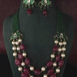 Puede incluir: Un conjunto de collar y pendientes a juego. El collar presenta cuentas de color burdeos y perlas de color crema. Los pendientes y los adornos del collar tienen diseños florales verdes y dorados. El collar se exhibe en un maniquí verde oscuro.