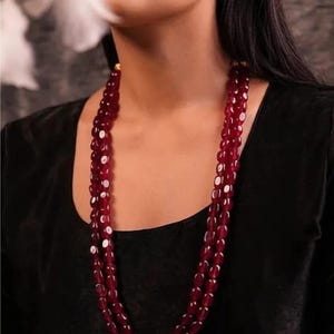 Op de afbeelding: Een ketting met drie strengen met ovale, dieprode kralen. De kralen zijn aan elkaar geregen, waardoor een gelaagd effect ontstaat. De ketting wordt gedragen op een zwarte top, waardoor de rijke kleur van de kralen wordt benadrukt.