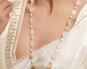 May include: A long necklace featuring alternating pink gemstones and small pearls, connected by gold-colored links. The necklace includes ornate gold pendants with red and pearl accents. The jewelry is displayed against a cream-colored fabric.