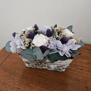 Peut inclure: Composition florale dans un contenant rectangulaire argenté et texturé. Elle comprend des roses blanches, des hortensias lavande, des fleurs violettes en forme de chardon et des feuilles d'eucalyptus vertes. Le contenant est posé sur une surface en bois.