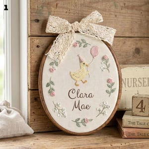 Puede incluir: Bastidor bordado ovalado con un pato con gorro de fiesta y un globo rosa. El nombre "Clara Mae" está bordado debajo. El diseño incluye bordados florales y un lazo de encaje, con marco de madera.