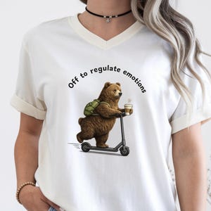 May include: Cream-colored V-neck t-shirt featuring a brown bear riding a black scooter, wearing a green backpack, and holding a coffee cup. The text "Off to regulate emotions" is arched above the bear. The shirt has rolled-up sleeves.