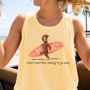 May include: A pale yellow sleeveless tank top featuring a graphic illustration of a brown monkey carrying a pink surfboard with flower and sun patterns. Below the illustration, the text reads, I don't want this feeling to go away.